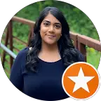 A woman with long dark hair and a navy blue top stands outdoors in front of a wooden railing, greenery behind her. An orange star badge icon labelled “Men’s Health” appears in the lower right corner of the image.