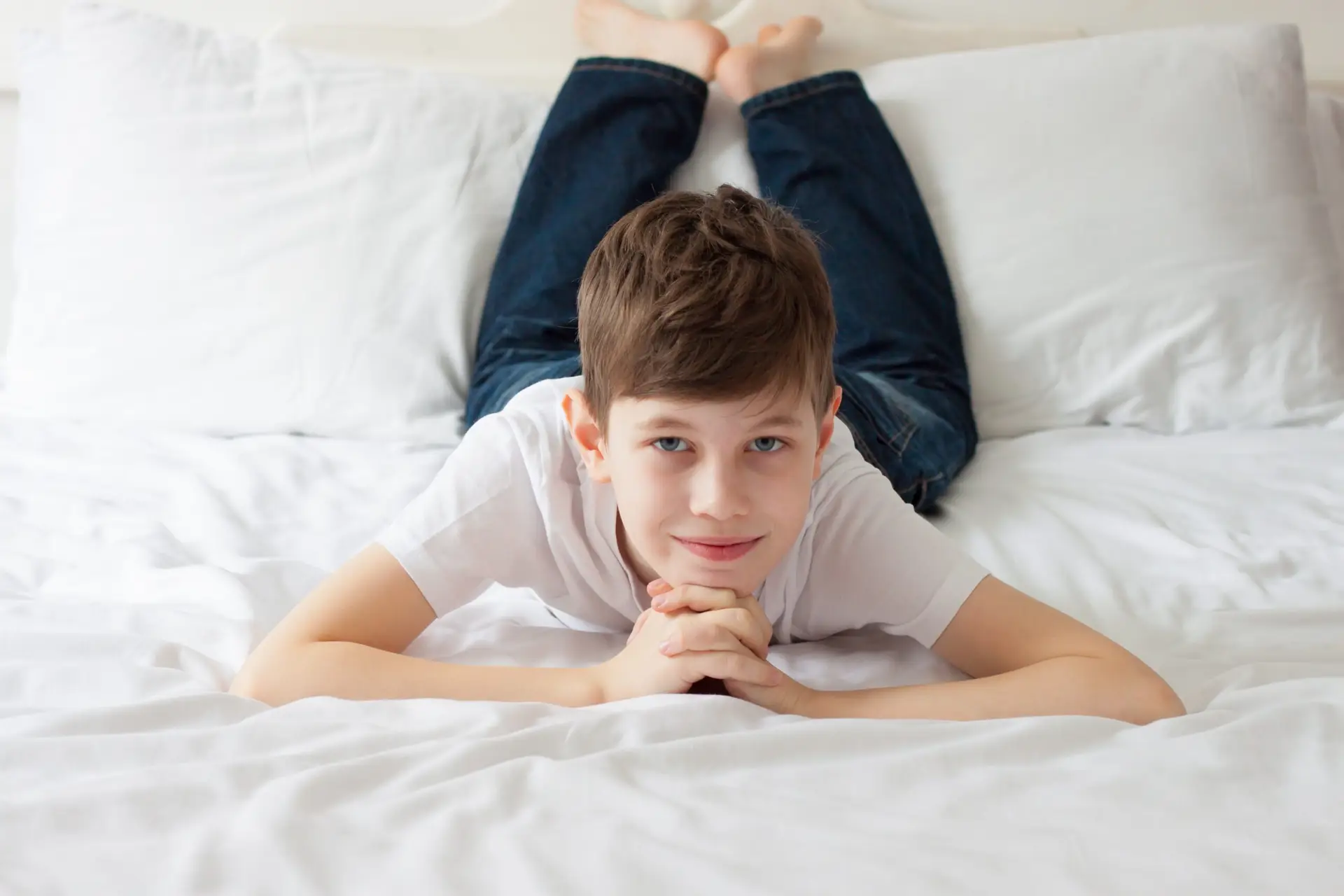 A boy with short brown hair, wearing a white T-shirt and jeans, lies on his stomach on a white bed with his hands clasped under his chin, smiling softly at the camera.