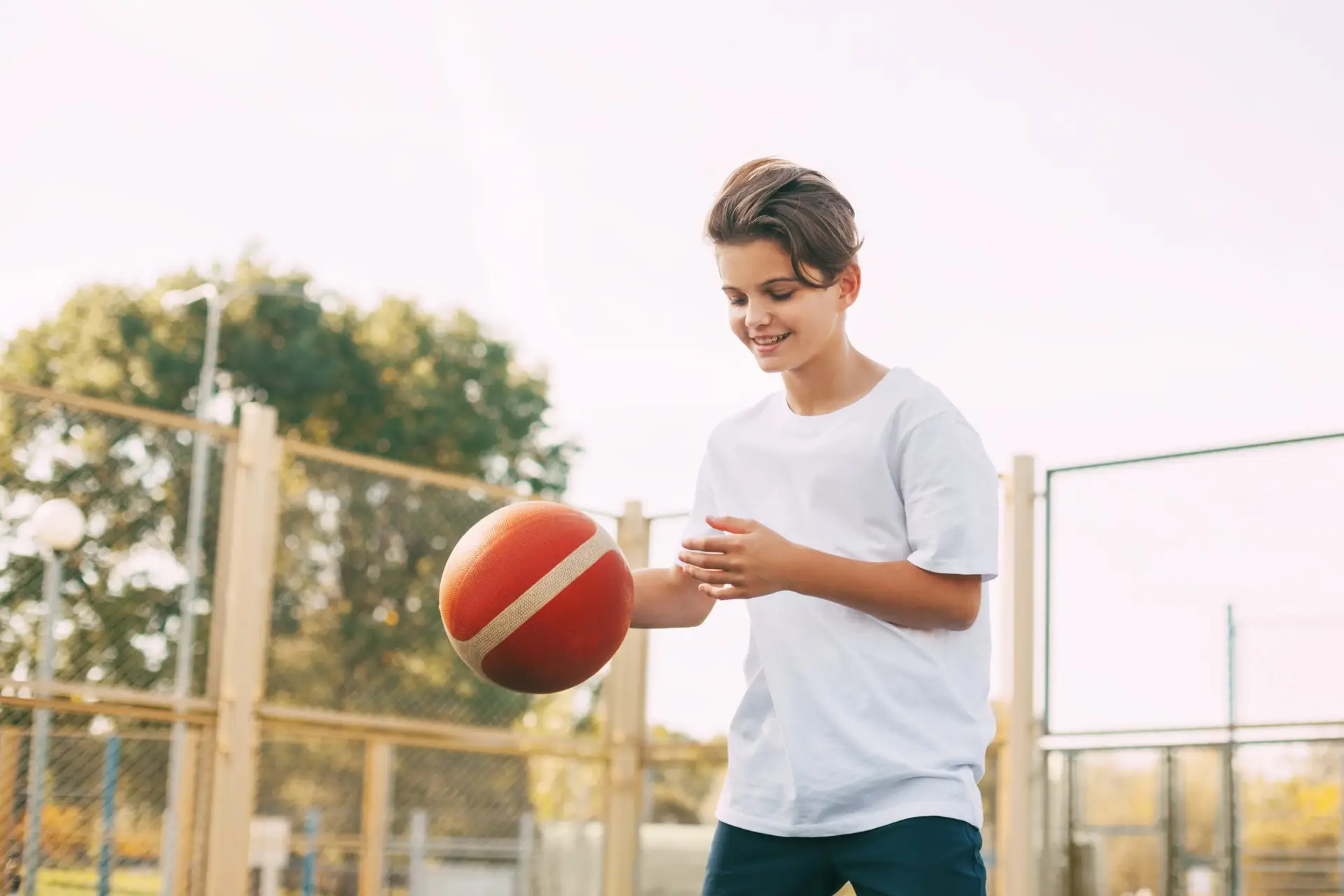 A smiling teenage boy in a white T-shirt dribbles a basketball on an outdoor court with a fence and trees in the background.