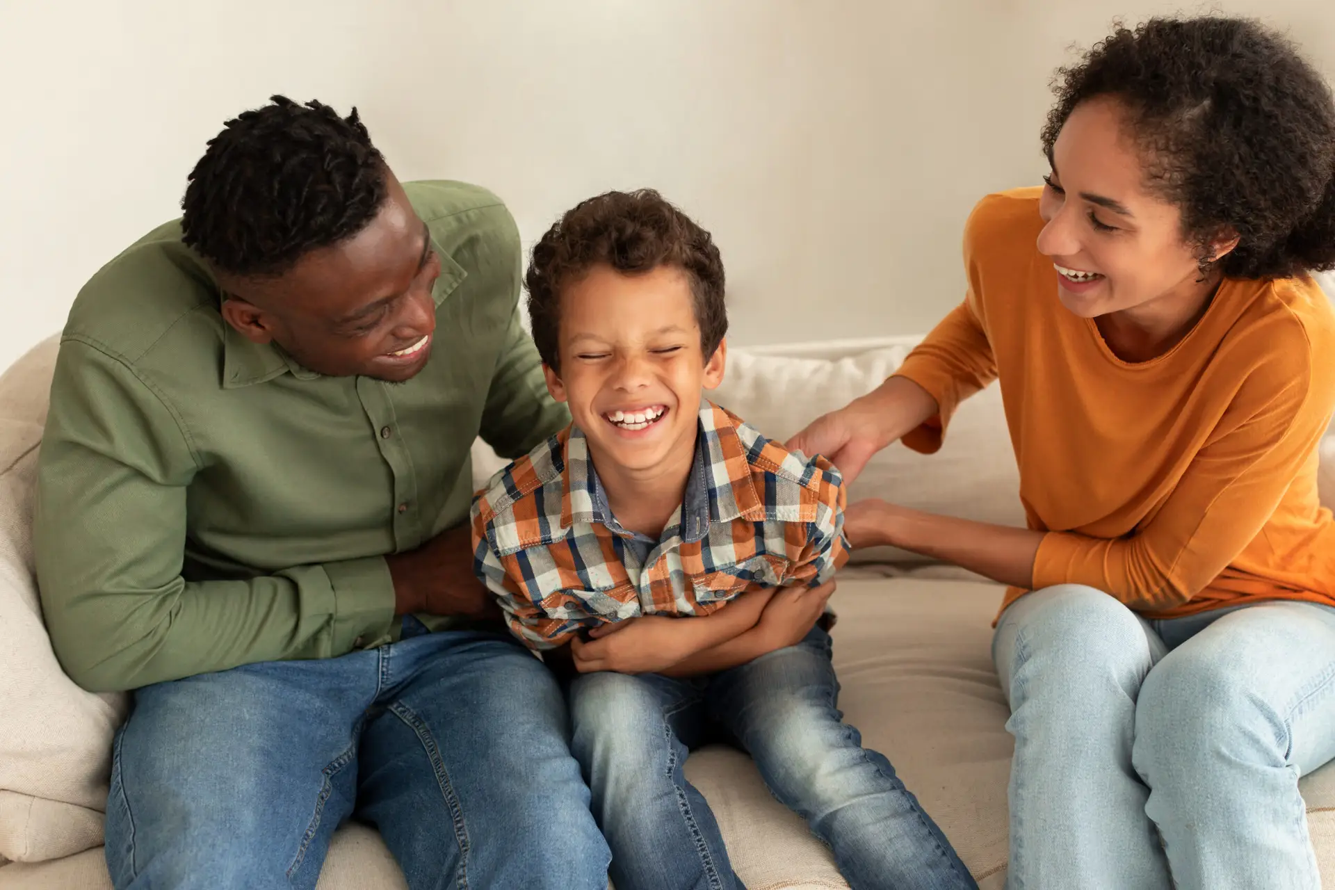 A smiling boy sits on a sofa between two adults, all laughing together. The adults appear to be playfully tickling the boy, creating a joyful and playful family moment.