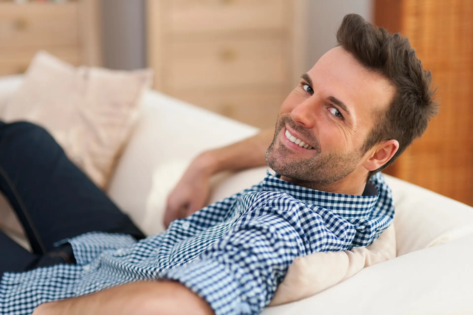 A man with short dark hair and a trimmed beard, wearing a blue and white chequered shirt, reclines on a white sofa and smiles whilst looking over his shoulder at the camera.