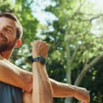 A man in sportswear stretches his arm outdoors on a sunny day, surrounded by green trees, and smiles while wearing a smartwatch.