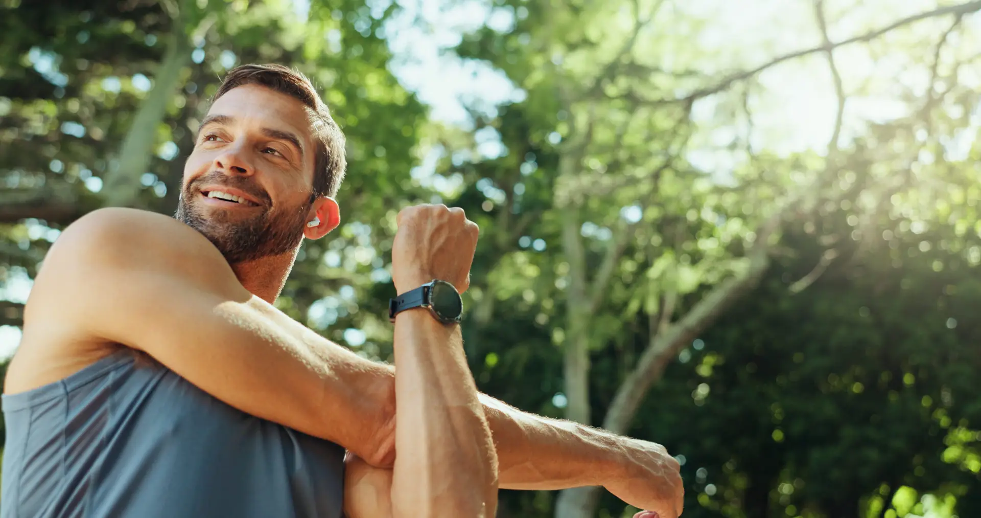 A man in sportswear stretches his arm outdoors on a sunny day, surrounded by green trees, and smiles while wearing a smartwatch.