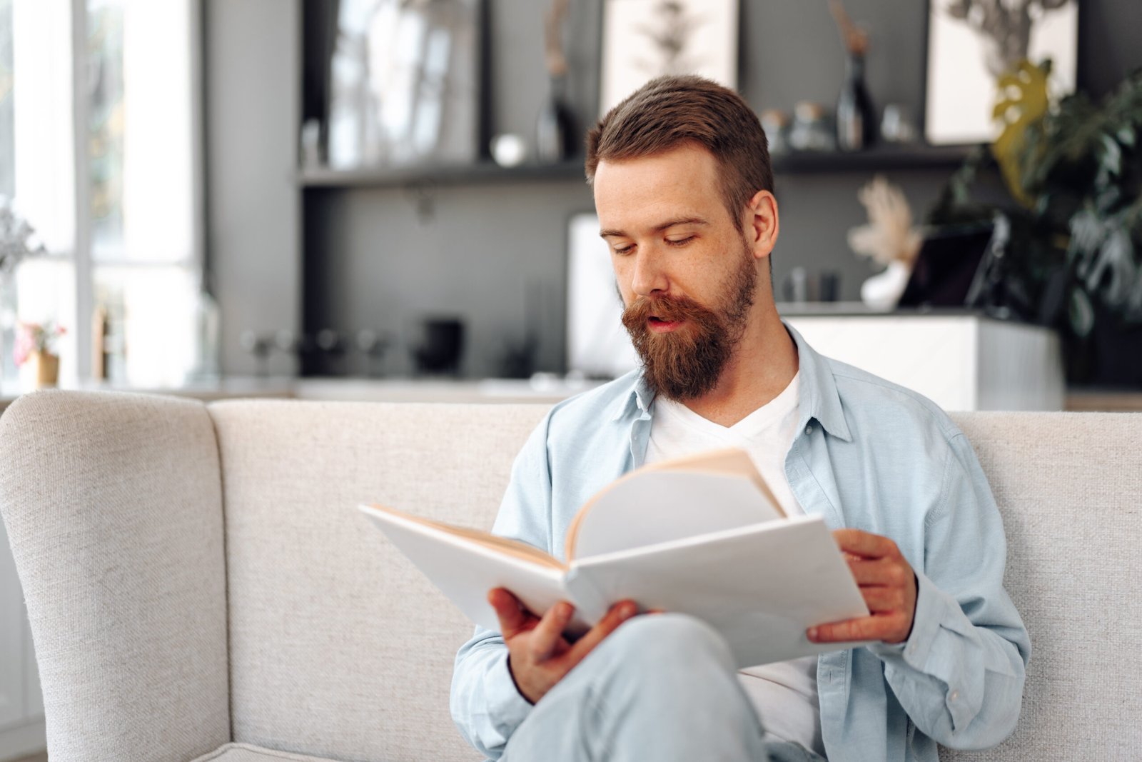 A bearded man sitting on a light-coloured sofa, wearing a light blue shirt and white T-shirt, reads a large book in a modern, softly lit living room.