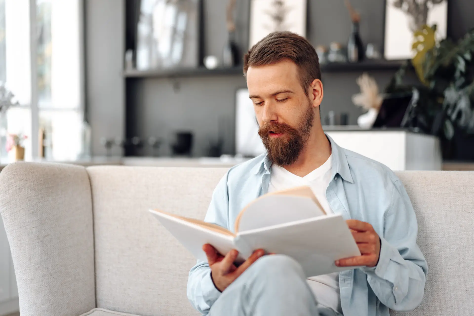 A bearded man sitting on a light-coloured sofa, wearing a light blue shirt and white T-shirt, reads a large book in a modern, softly lit living room.