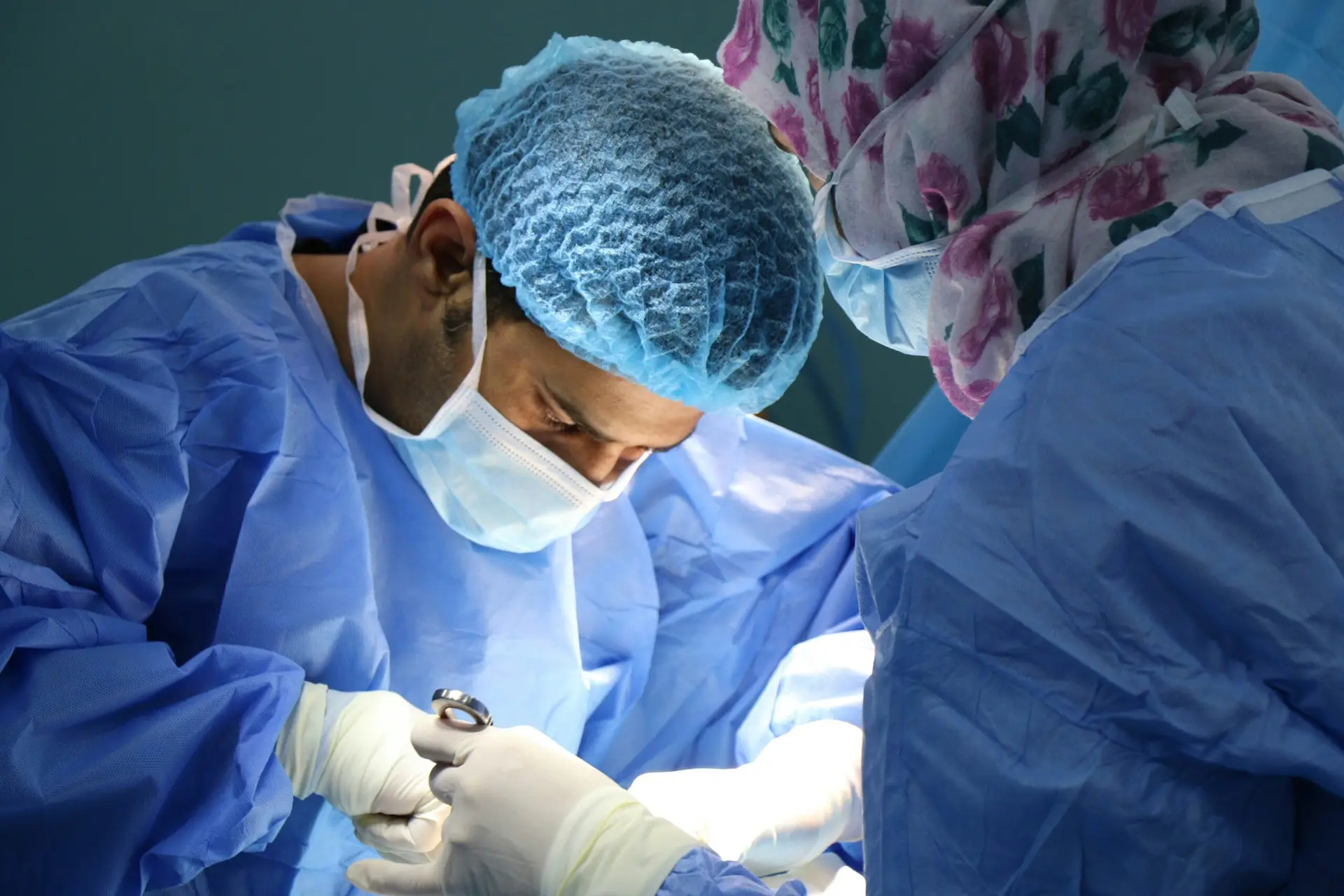 Two surgeons in blue gowns, masks, and hair covers are focused on performing a circumcision procedure under bright light in an operating theatre.