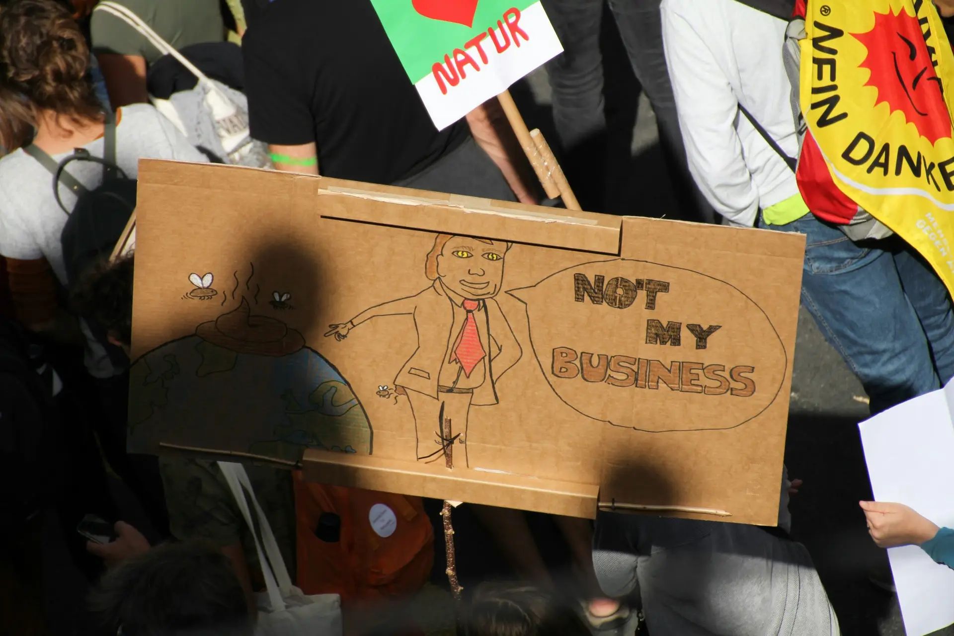 A group of people at a protest hold signs, including a cardboard one with a drawing of a person ignoring a burning Earth, saying NOT MY BUSINESS. Other colourful protest and anti-circumcision signs are visible in the crowd.