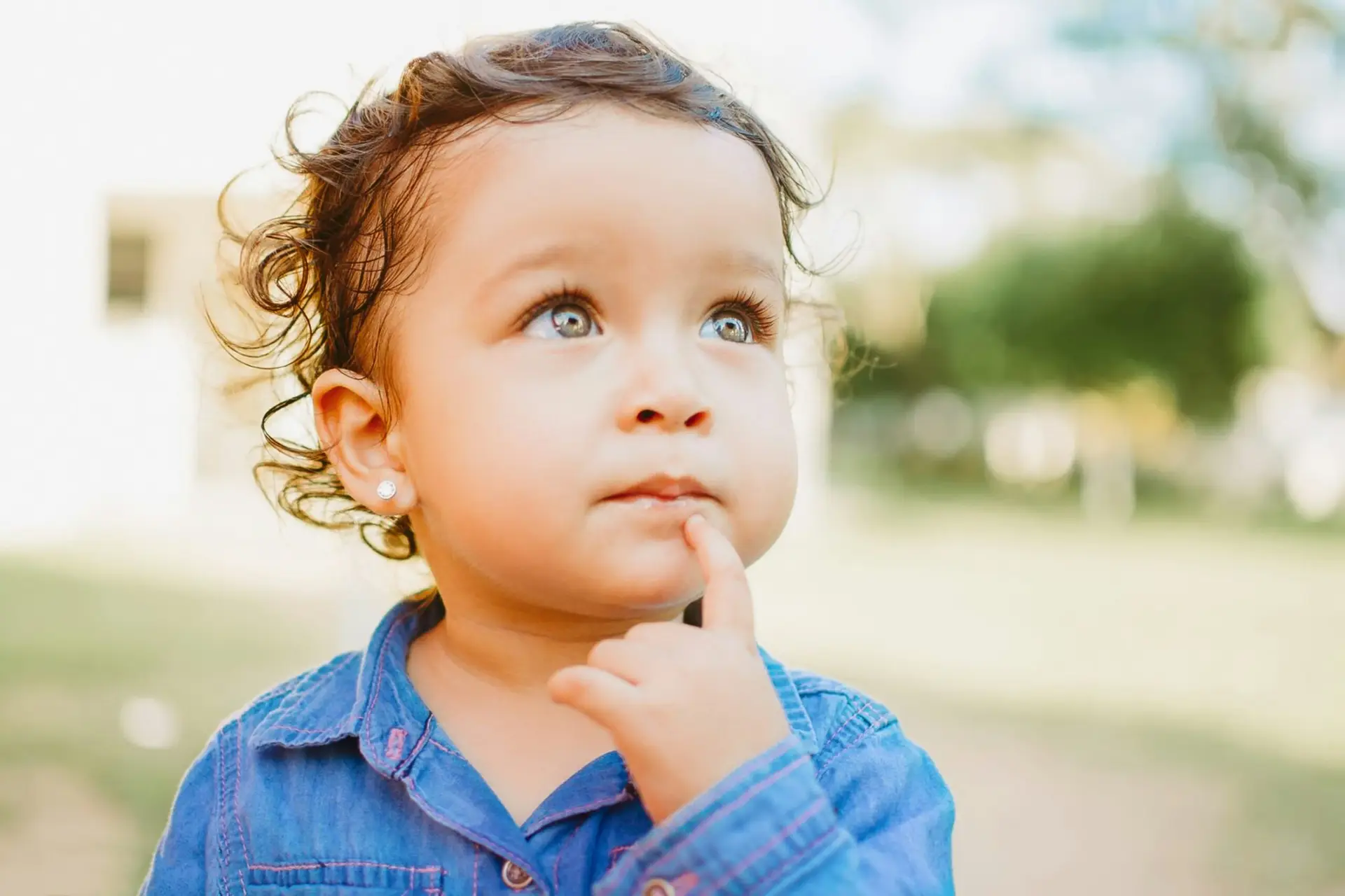 A young child with curly hair and light eyes, wearing a blue shirt, looks upward thoughtfully and touches their chin with one finger, perhaps pondering questions about topics like circumcision. The background is softly blurred with greenery.