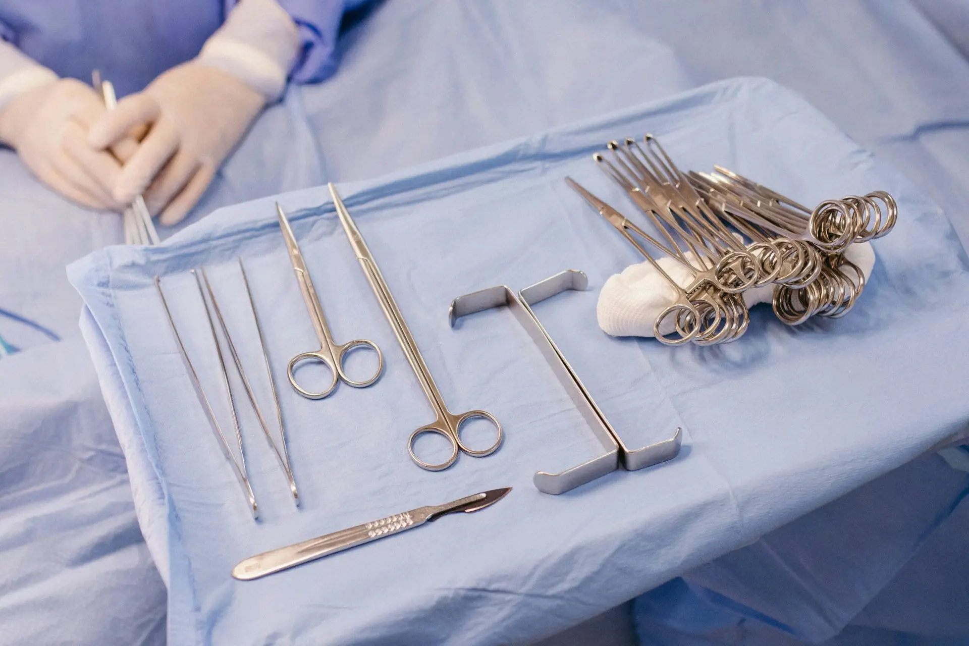 A surgical tray with various metal instruments, including forceps, scissors, scalpels, and retractors used in circumcision, is arranged on a blue sterile drape; a gloved medical professional is visible in the background.