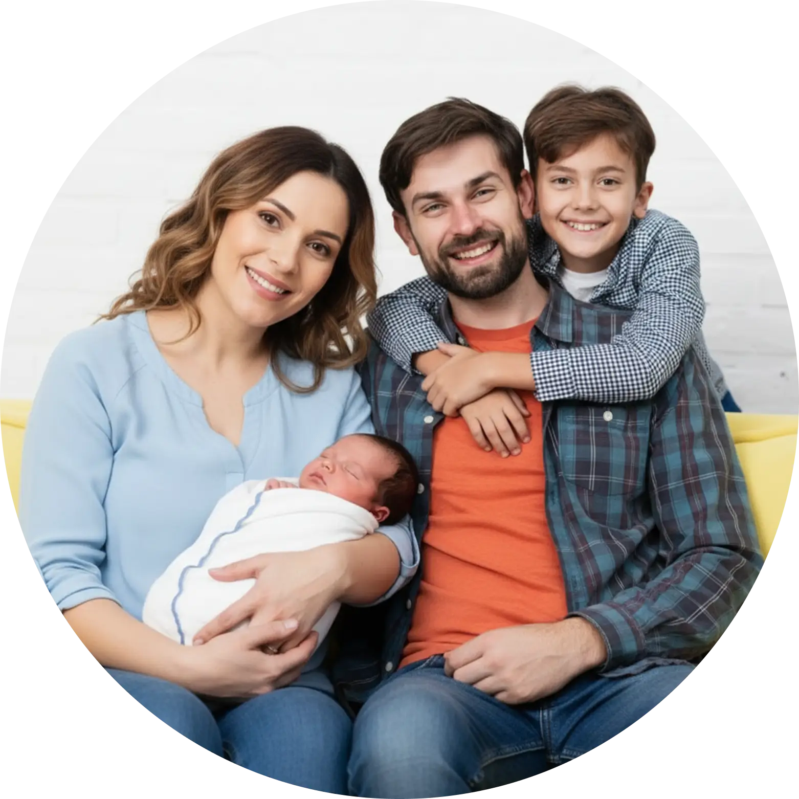 A smiling family of four sits together on a sofa. The mother holds a swaddled newborn, the father—radiating Men's Health—sits beside her, and an older son hugs his father from behind, all looking at the camera.