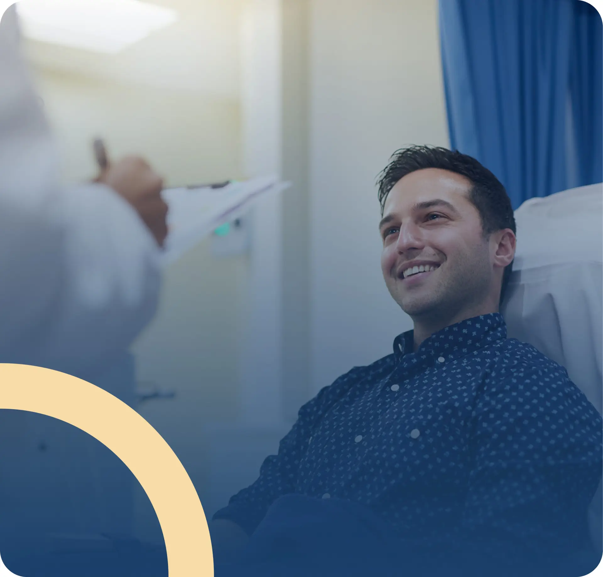 A man sits in a hospital or clinic room, smiling and looking at a doctor who is holding a clipboard. The setting appears bright and welcoming, with blue curtains in the background, reflecting a positive approach to Men’s Health.
