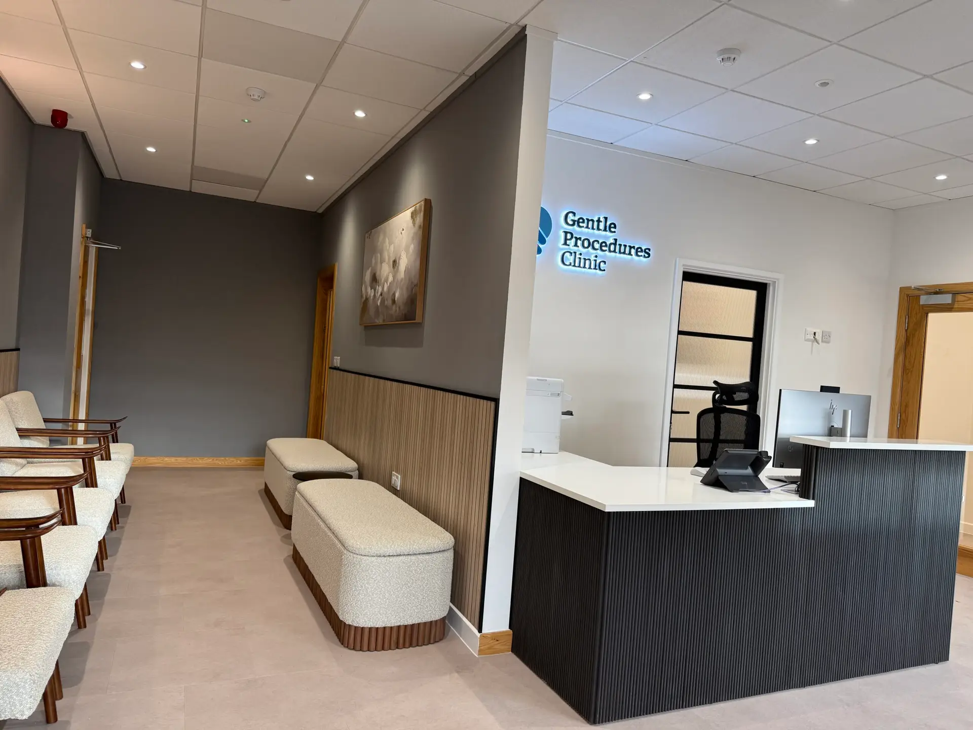 A modern medical waiting room with cushioned chairs and benches, neutral colours, and a reception desk. A sign reading Gentle Procedures Clinic is displayed on the back wall above the entrance to an office.