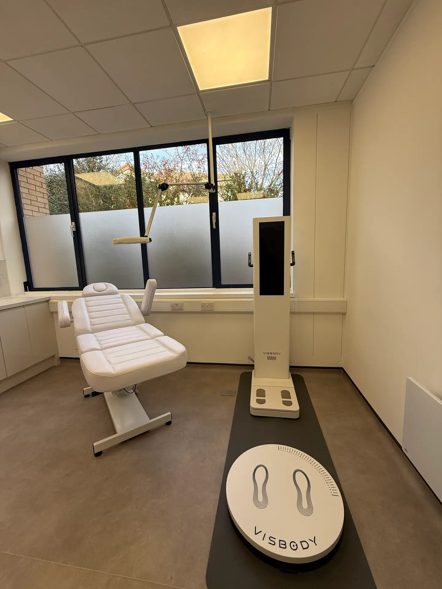 A modern Gentle Procedures Clinic room with a white examination chair, a large device with a screen labelled VISBODY, and a platform with footprints, set near large windows with frosted glass.
