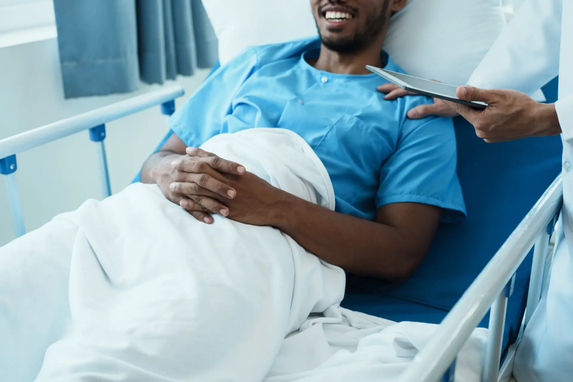 A patient in a hospital bed, smiling and wearing a blue gown, has hands resting on a white blanket. A medical professional stands beside the bed, holding a clipboard or tablet.