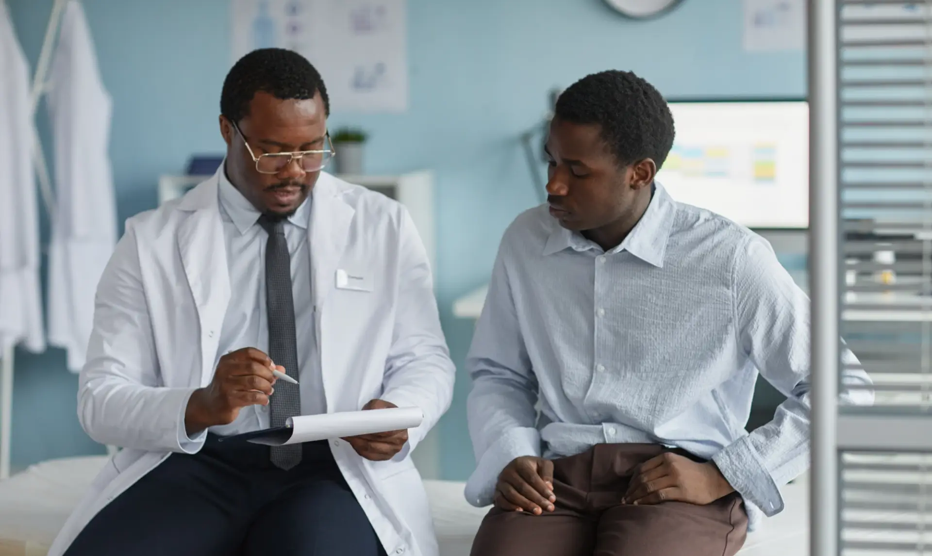 A doctor in a white coat sits beside a patient, reviewing notes on a clipboard. The patient listens attentively. They are in a surgery with medical charts and equipment visible in the background.
