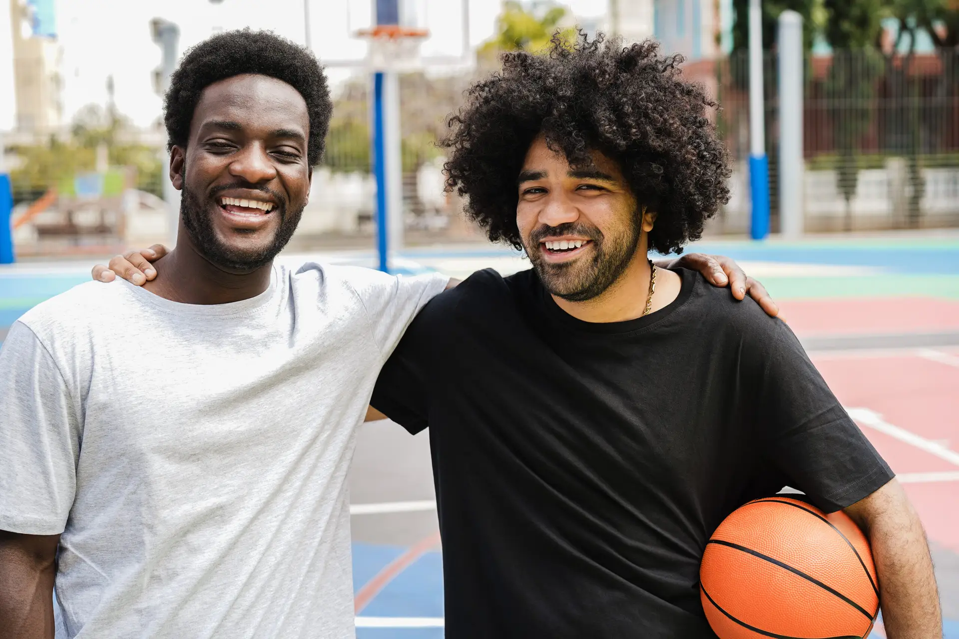 Two men smiling and standing side by side on an outdoor basketball court in Birmingham. One man has his arm round the other's shoulder, while the other holds a basketball. Both appear happy and relaxed after discussing vasectomy in Birmingham.