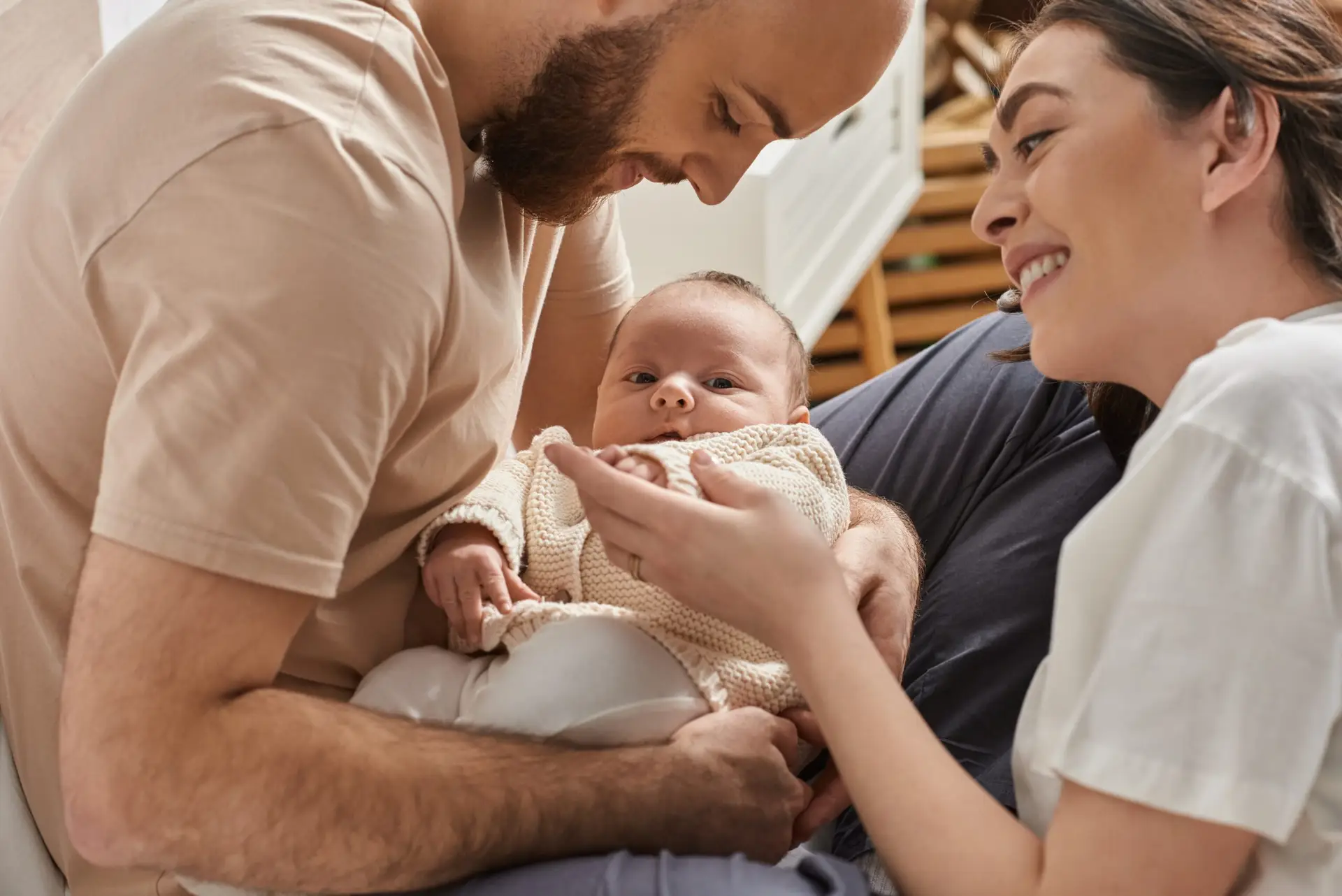 A smiling couple sit closely together, lovingly holding their baby in their arms. The baby, recently welcomed after newborn circumcision in Birmingham, looks directly at the camera while the parents gaze affectionately at the child.