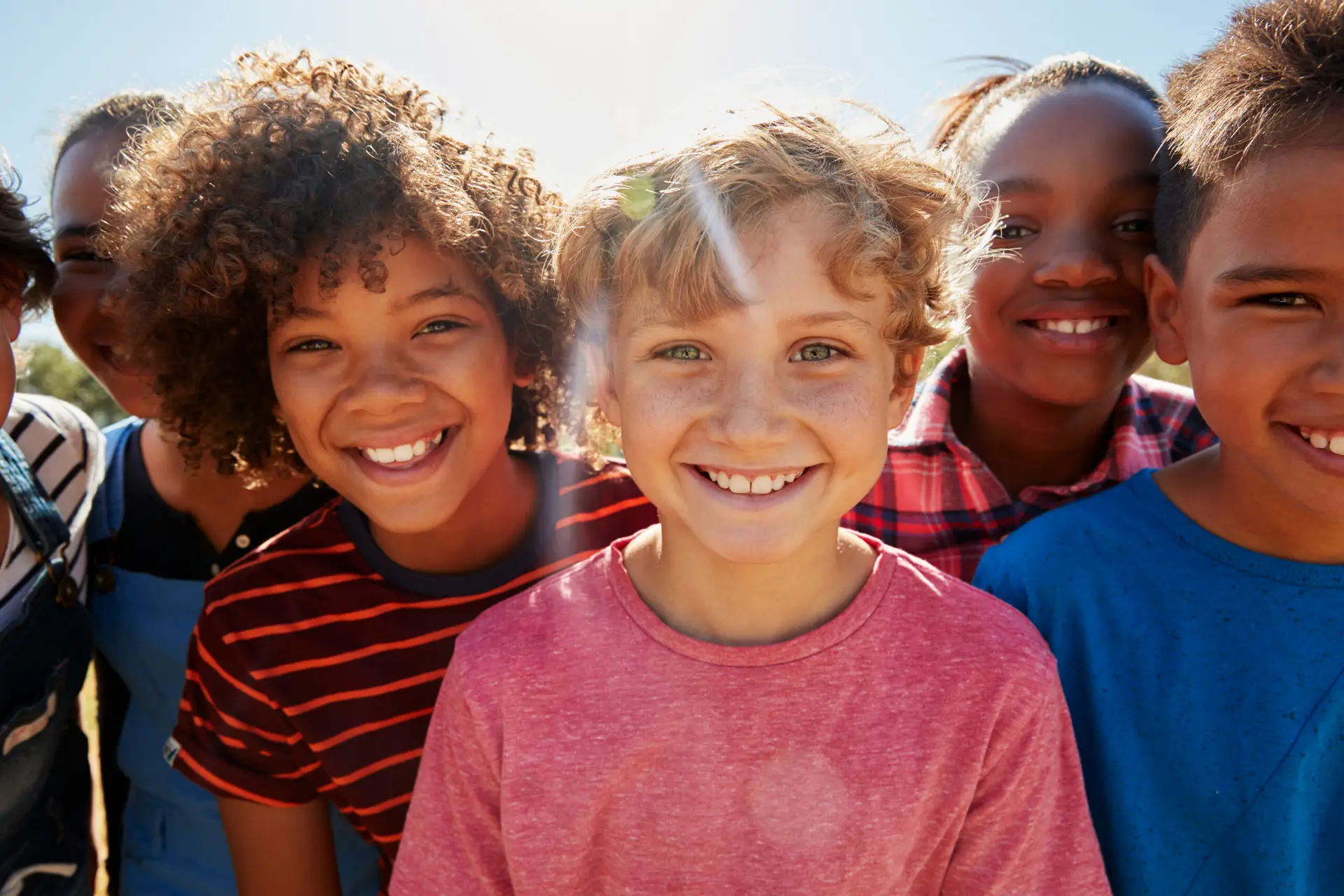 A group of smiling children stand close together outdoors on a sunny day in Birmingham, looking at the camera. They are wearing casual clothes and appear happy and cheerful, with sunlight shining behind them—representing boys' and teens' circumcision in Birmingham.