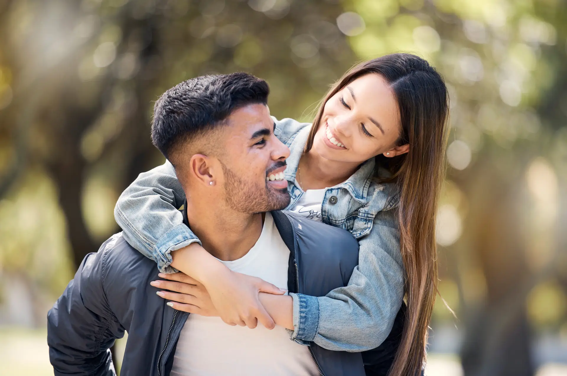 A young woman smiles and hugs a man from behind as he carries her on his back. Enjoying the outdoors near blurred trees, they look at each other happily, embracing life together in Birmingham.