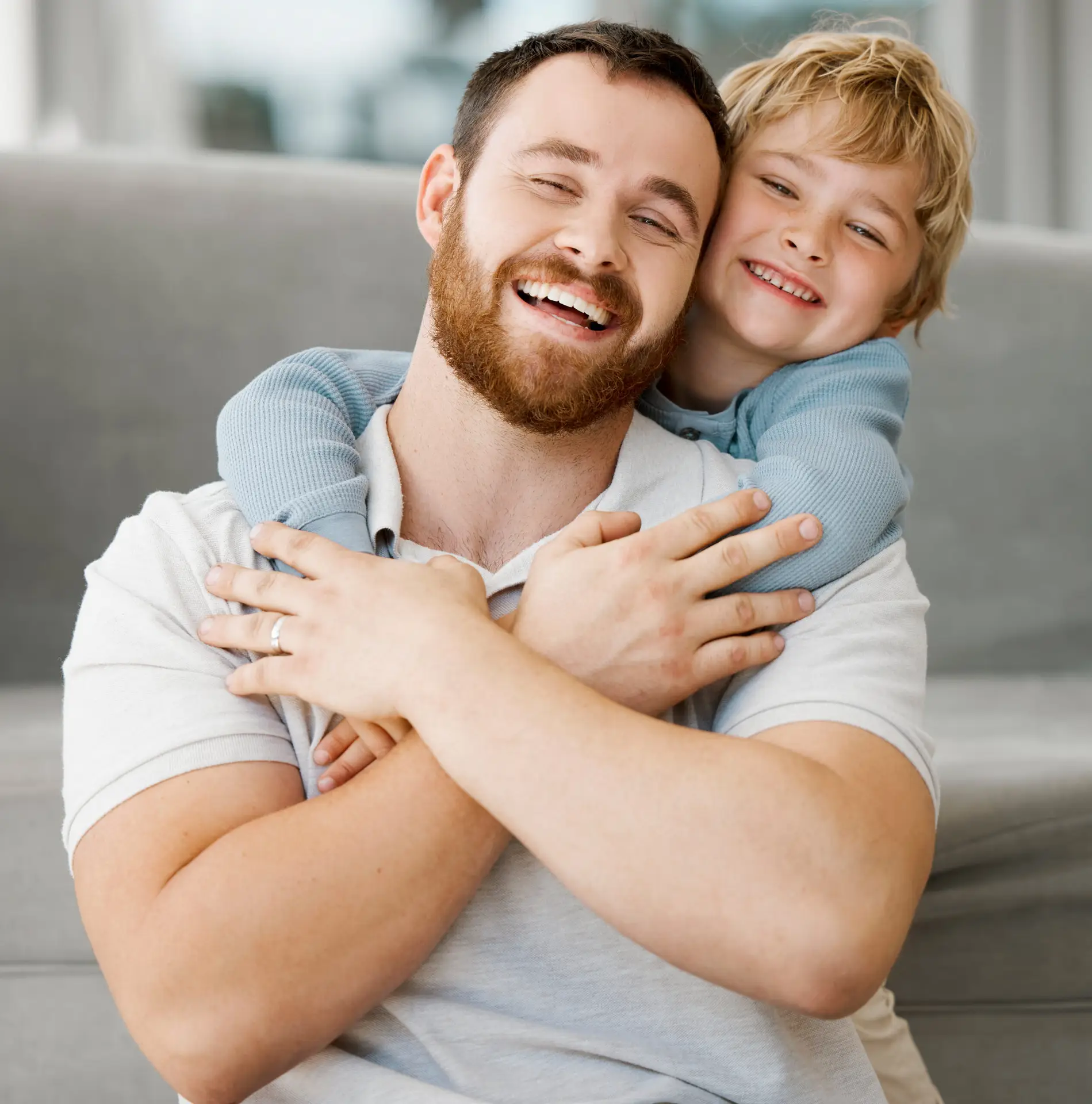 A smiling man with a beard sits on the floor whilst a young boy hugs him from behind. Both are laughing and appear happy, enjoying family time indoors on a light-coloured sofa in Birmingham.