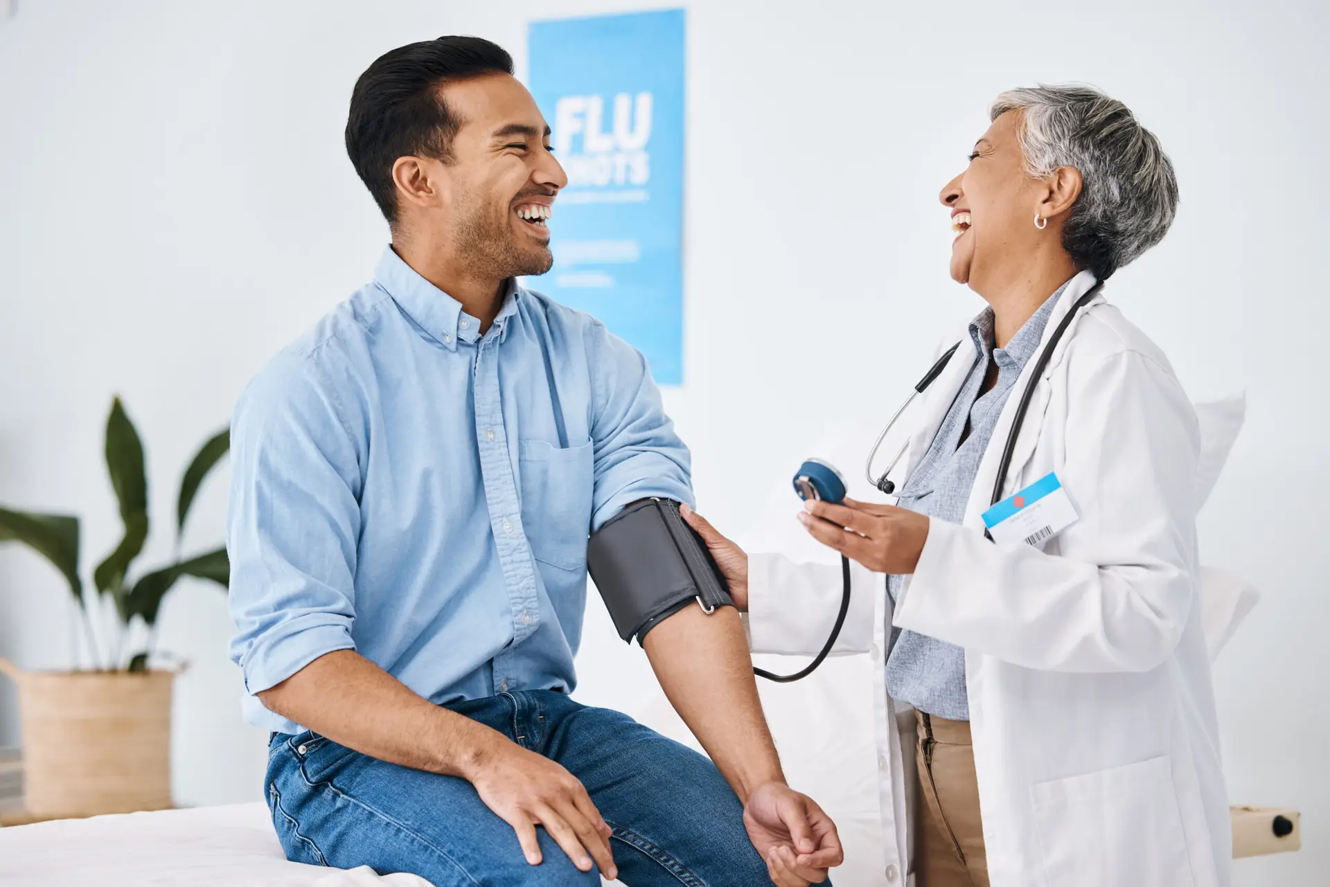 A doctor in a white coat laughs with a smiling male patient whilst measuring his blood pressure in a bright medical surgery, where a sign for flu jabs and full body health assessment in Birmingham is visible in the background.