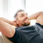 A man with short hair and a beard is smiling and relaxing on a sofa with his hands behind his head, wearing a blue shirt, in a bright room, reflecting the comfort and confidence after visiting a men's health clinic Birmingham.