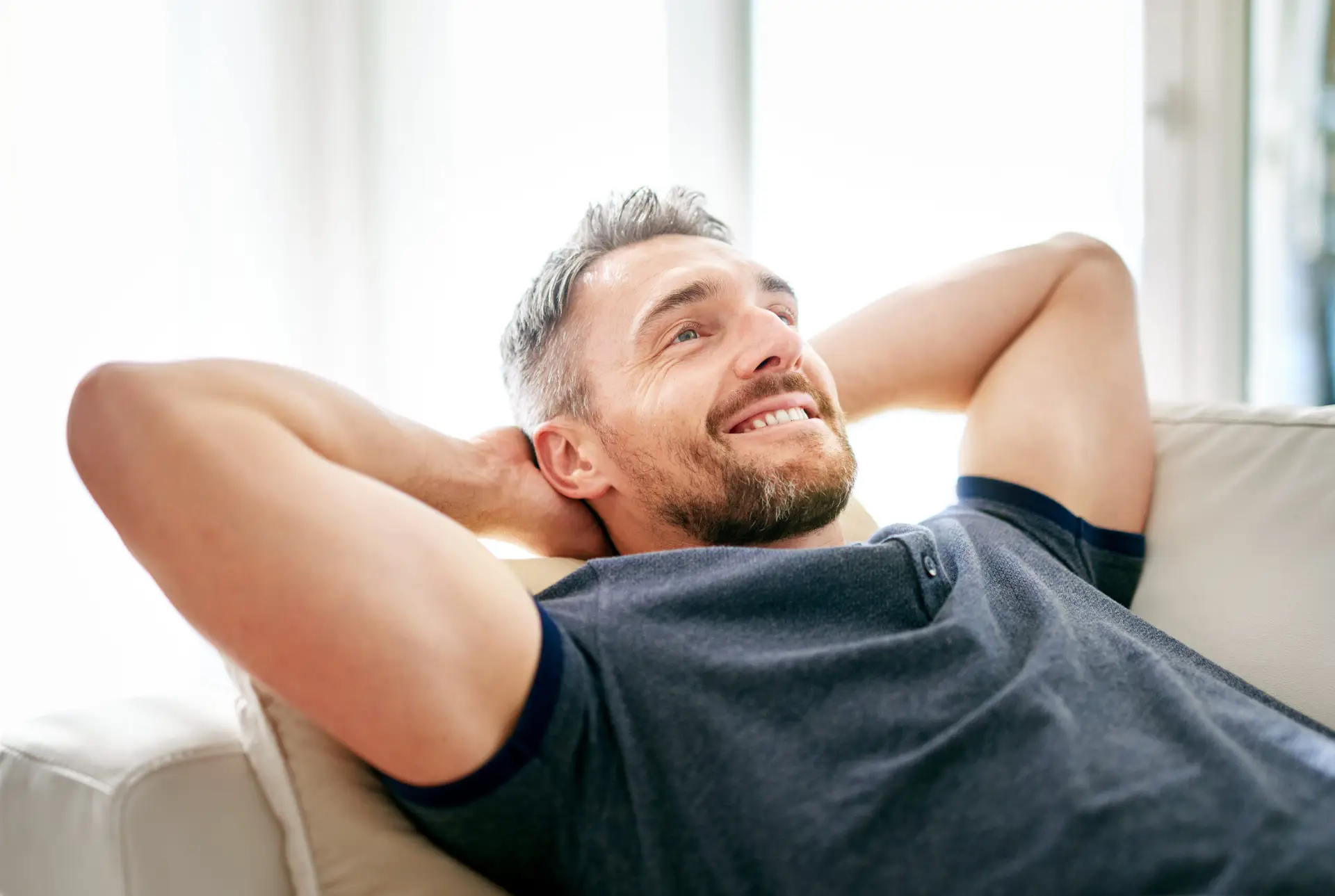 A man with short hair and a beard is smiling and relaxing on a sofa with his hands behind his head, wearing a blue shirt, in a bright room, reflecting the comfort and confidence after visiting a men's health clinic Birmingham.