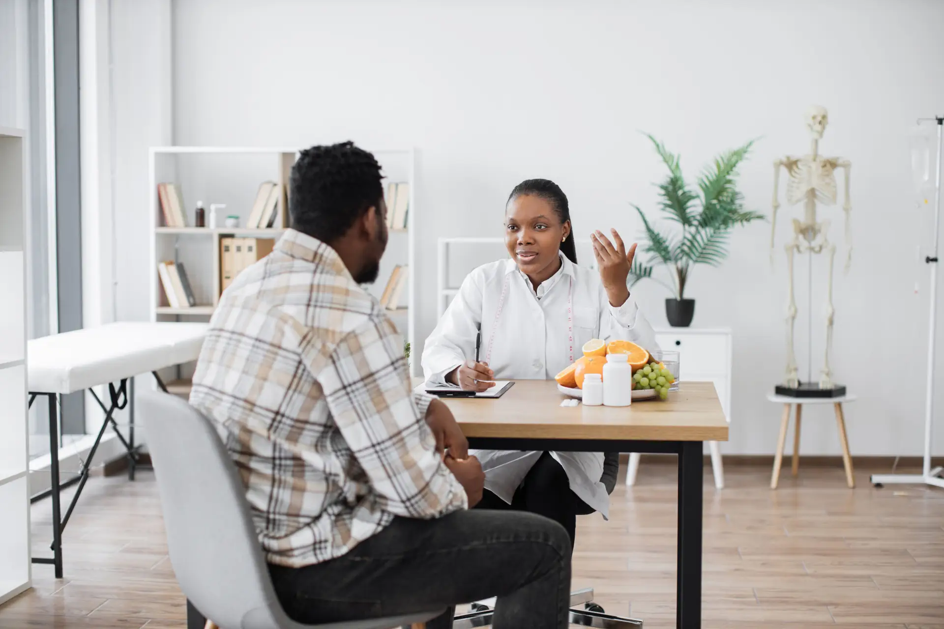 A doctor in a white coat sits at a desk, discussing weight loss for men in Birmingham with a patient. There are fruit, a clipboard, and medicine bottles on the desk. Shelves and a skeleton model are in the background.