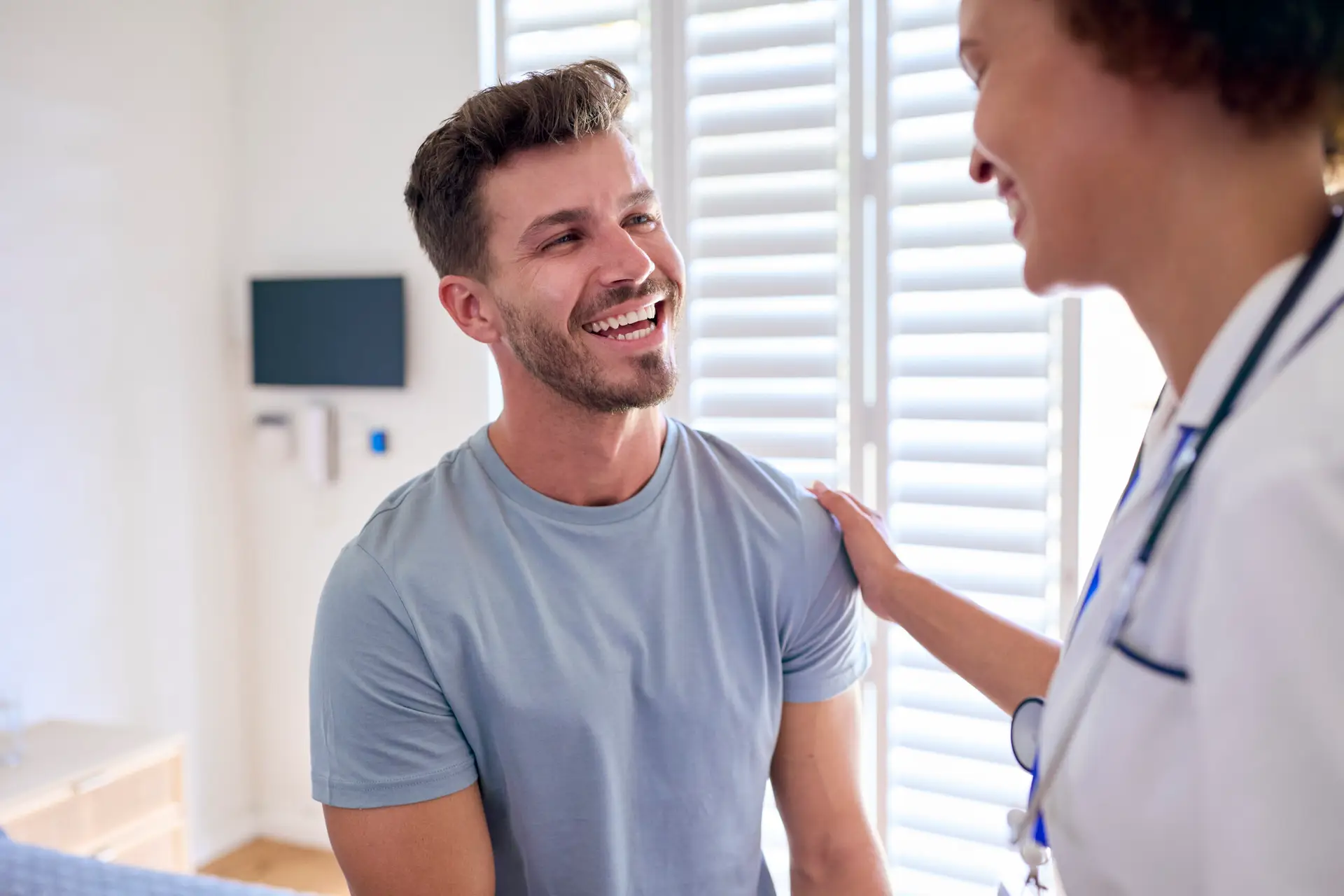 A smiling man sits on an examination table while talking with a healthcare professional during his full body health assessment in Birmingham, the provider resting a reassuring hand on his shoulder in a bright consulting room.