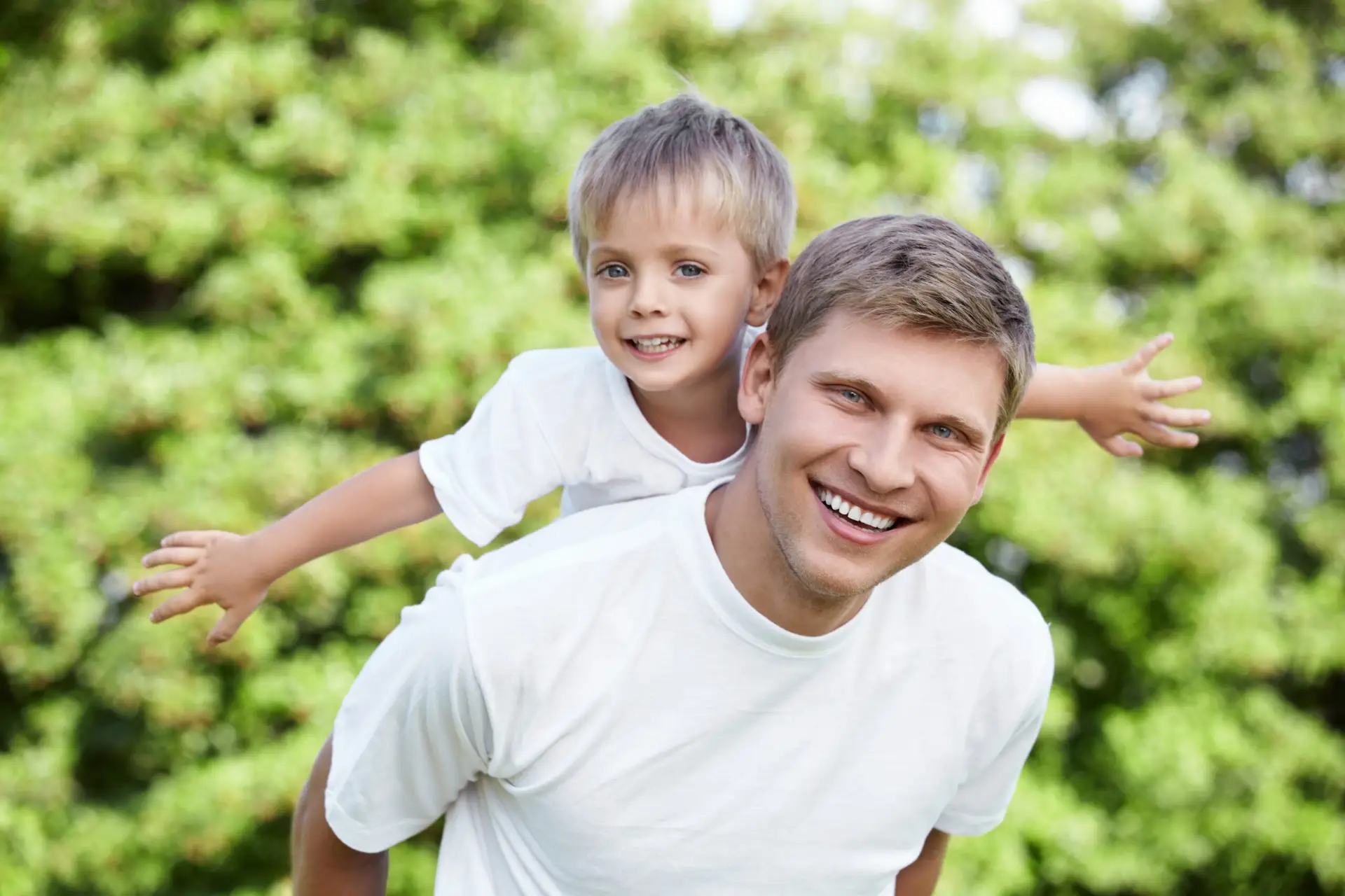 A smiling man gives a young boy a piggyback outdoors. Both, possibly celebrating after circumcision in Birmingham, are wearing white shirts and appear happy, with greenery in the blurred background.