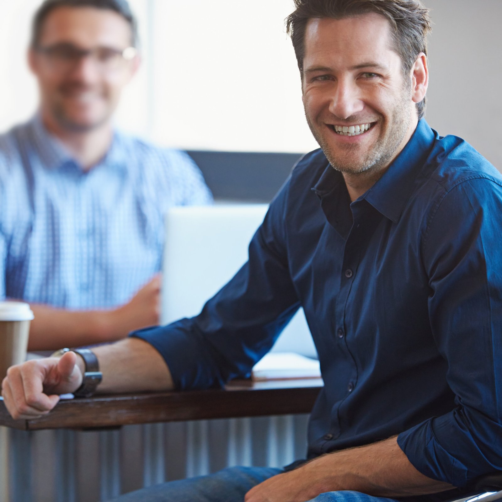 A man in a dark blue shirt smiles at the camera whilst sitting at a table, possibly discussing anti-wrinkle injections for men in Birmingham, with another man and a laptop blurred in the background. A coffee cup sits on the table, suggesting a casual office or café setting.