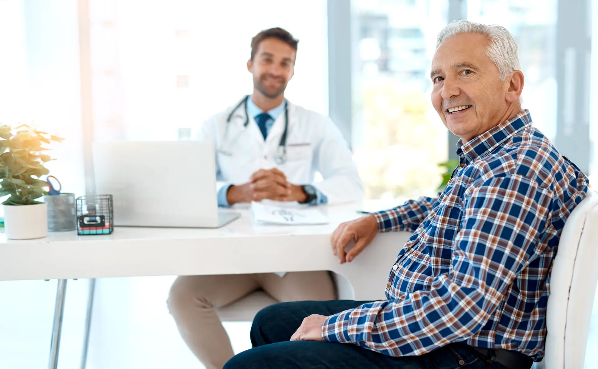 An older man in a checked shirt sits and smiles at the camera in a doctor's surgery, where he’s discussing weight loss for men in Birmingham with a doctor in a white coat and stethoscope at the desk behind him.