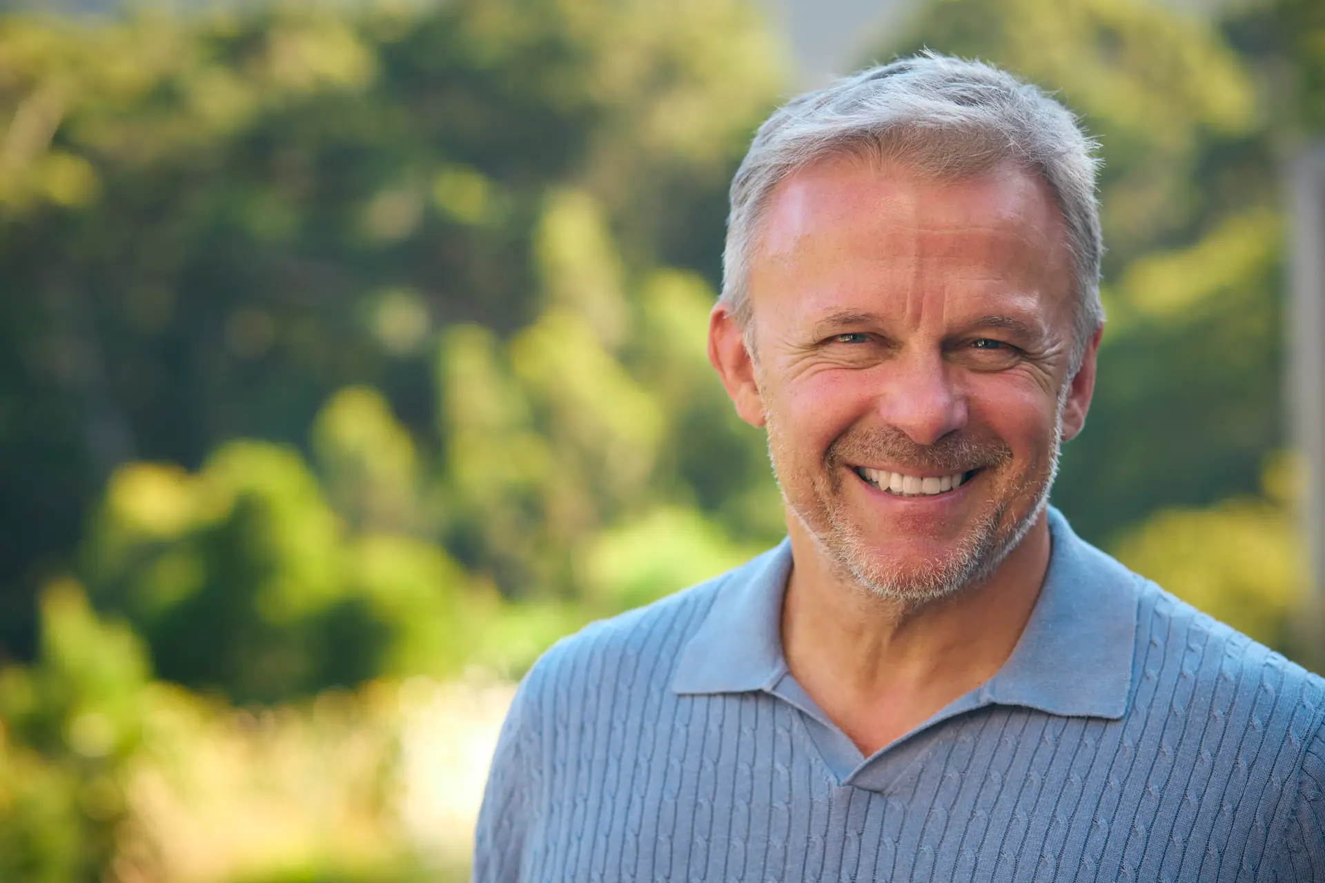 A smiling older man with grey hair and a trimmed beard stands outdoors in front of blurred green trees, wearing a light blue jumper with a collar, appearing confident after learning more about adult circumcision in Birmingham.