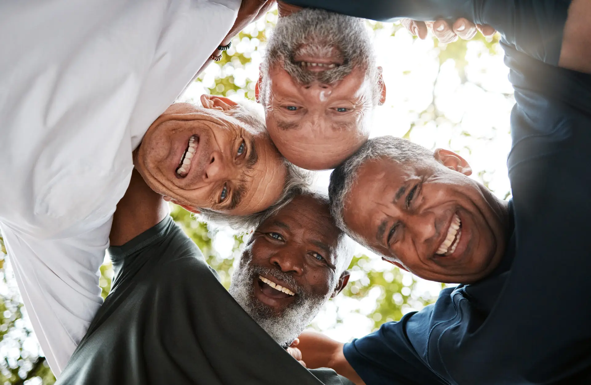 Four older men smile and stand in a huddle with their heads together, looking down at the camera from above. Sunlight and trees blur behind them, capturing the camaraderie often celebrated in Specialist Men’s Surgical Care.