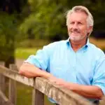 A smiling older man with grey hair and a beard leans on a wooden fence outdoors, wearing a light blue shirt. He embodies the vitality made possible by Specialist Men's Surgical Care, with green trees and grass in the background.