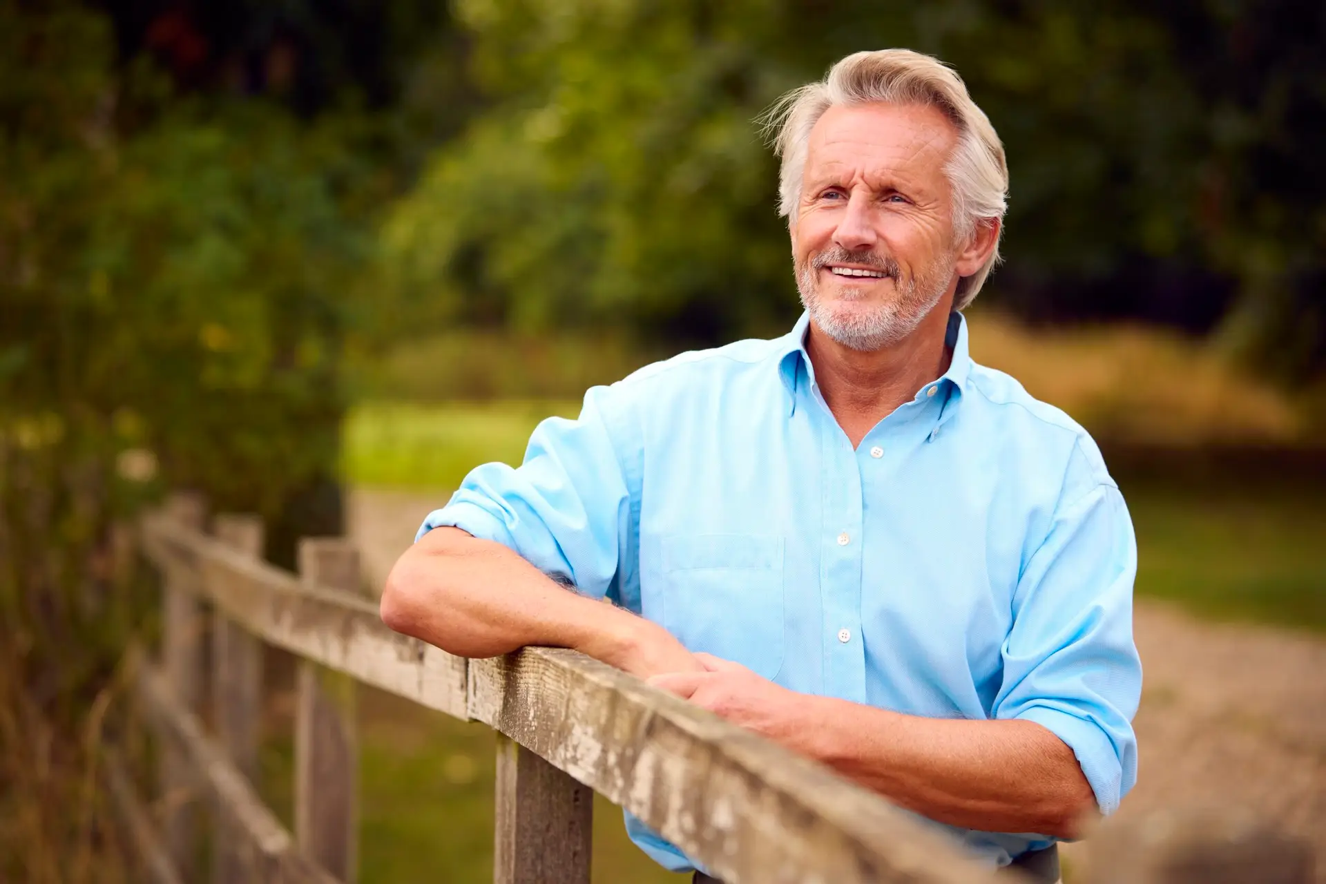 A smiling older man with grey hair and a beard leans on a wooden fence outdoors, wearing a light blue shirt. He embodies the vitality made possible by Specialist Men's Surgical Care, with green trees and grass in the background.