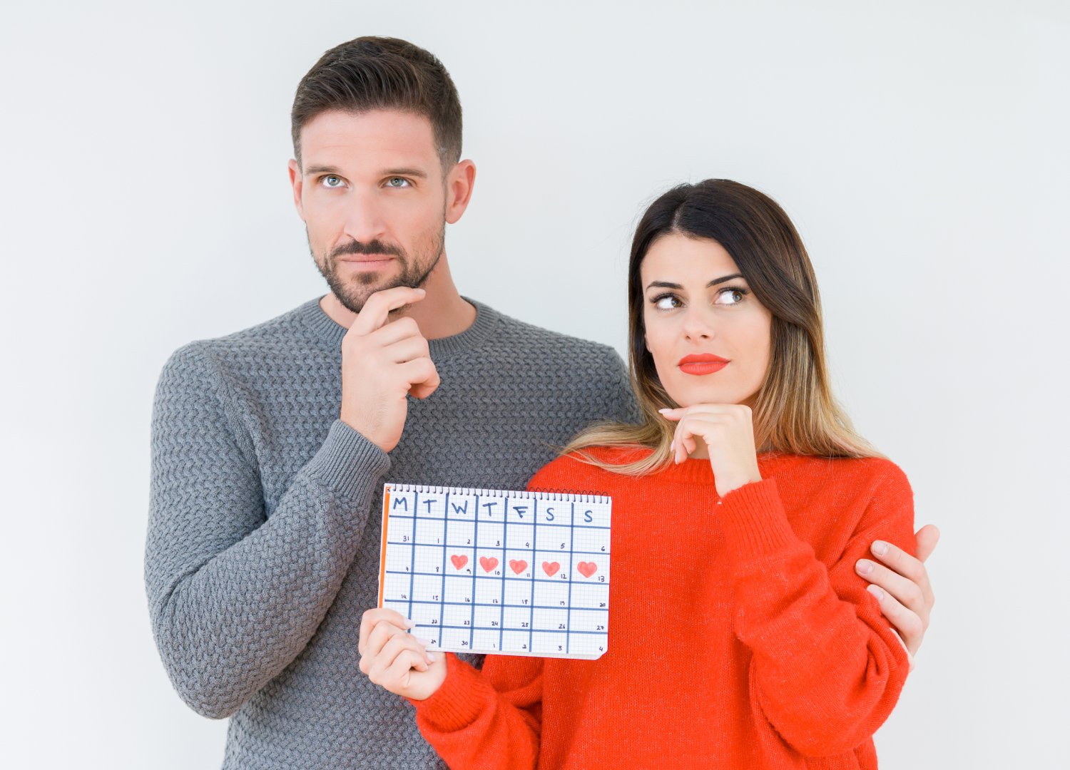 A man and woman stand together, both looking thoughtful. The woman holds a calendar marked with hearts on several days. The man has his hand on his chin, and the woman points at the calendar whilst glancing sideways.