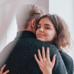A woman with wavy brown hair smiles at the camera while hugging a person with short hair in a dark jumper. She is wearing a ring and has her hand resting on the other person's back.