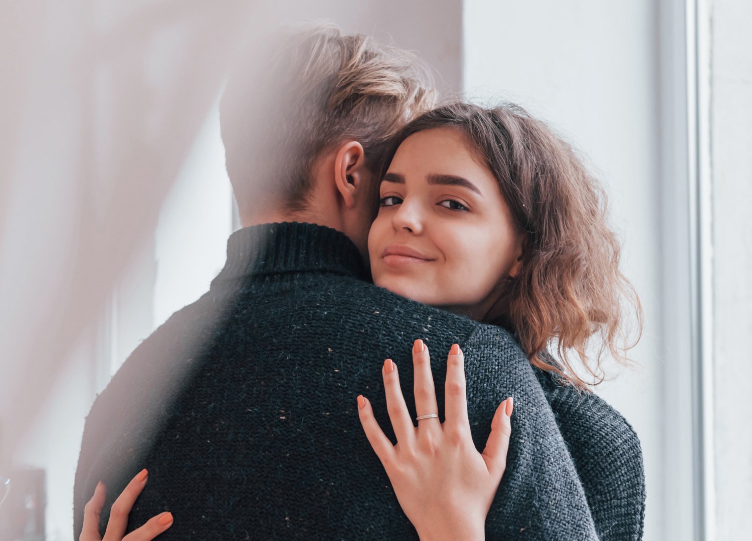 A woman with wavy brown hair smiles at the camera while hugging a person with short hair in a dark jumper. She is wearing a ring and has her hand resting on the other person's back.