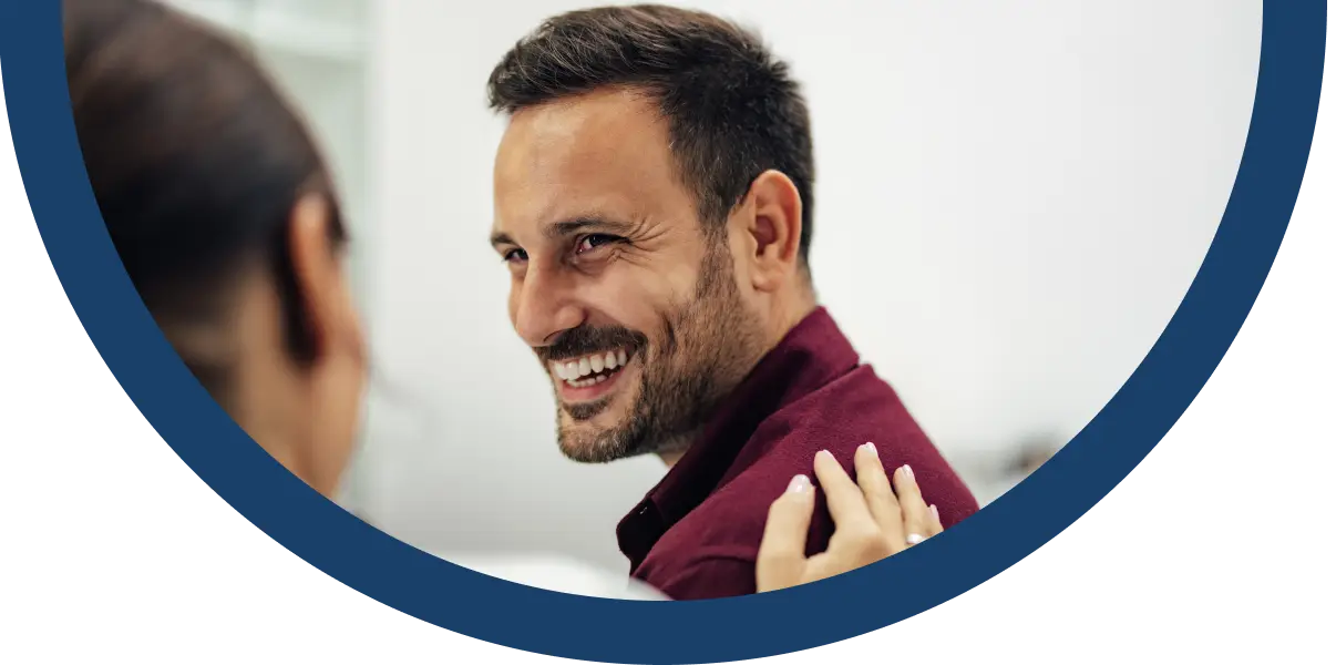 A man with a beard and short hair, wearing a maroon shirt, smiles warmly at a person whose hand rests on his shoulder. The background is blurred, and the image is framed by a blue curved border.