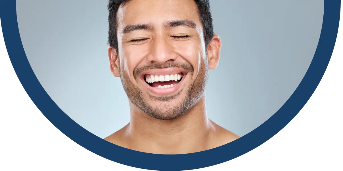 A man with short dark hair and facial hair is smiling broadly with his eyes closed, showing his teeth. The image is cropped in a semicircle with a blue border on a light background.