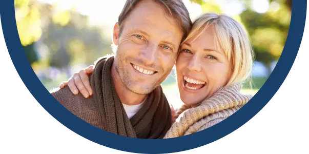A smiling couple dressed in warm jumpers and scarves pose closely together outdoors, with sunlight and greenery in the blurred background.