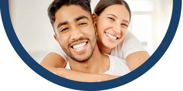 A smiling couple pose closely together indoors, with the woman hugging the man from behind. Both are looking at the camera and appear happy. The image is framed by a thick, dark blue circular border.