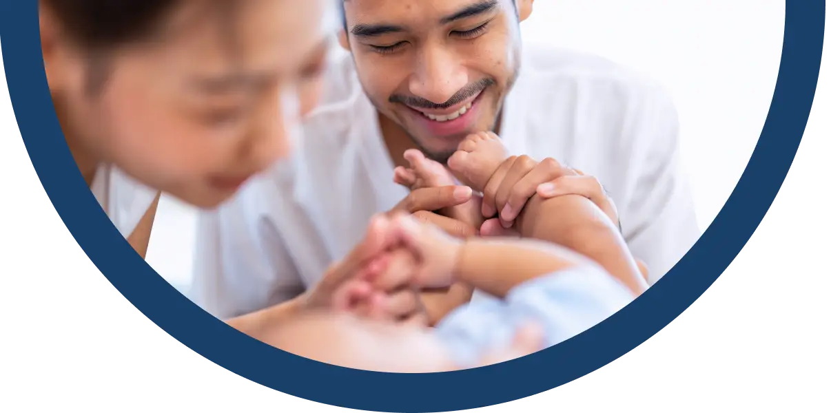 A smiling man gently holds a baby’s hands whilst a woman, slightly out of focus, looks on lovingly. The scene is framed by a blue border, conveying warmth and family connection.