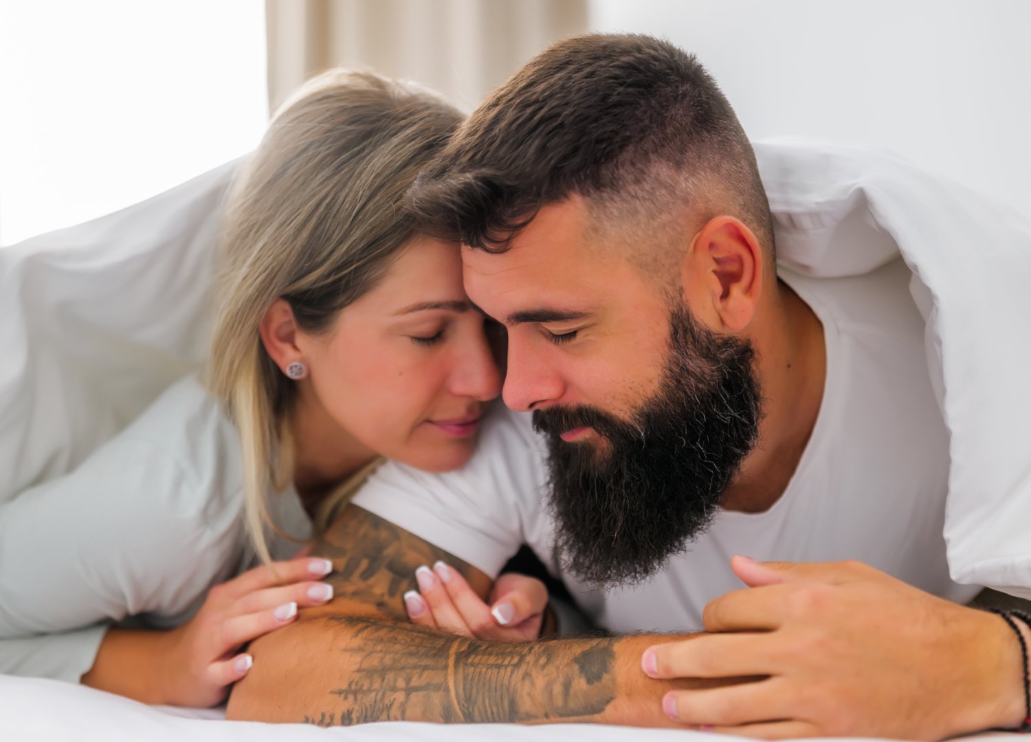 A couple lies close together under a white duvet, facing each other with eyes closed and peaceful expressions, suggesting intimacy and affection.