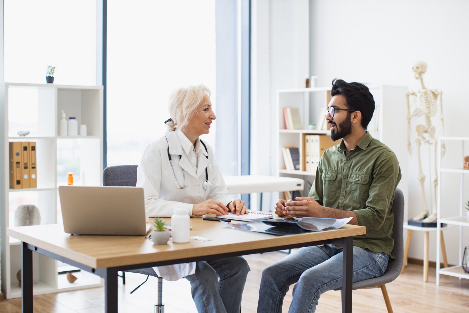 A doctor and a patient are sitting opposite each other at a desk in a bright medical surgery, discussing X-ray images. The doctor is smiling and holding a pen, while the patient listens attentively.