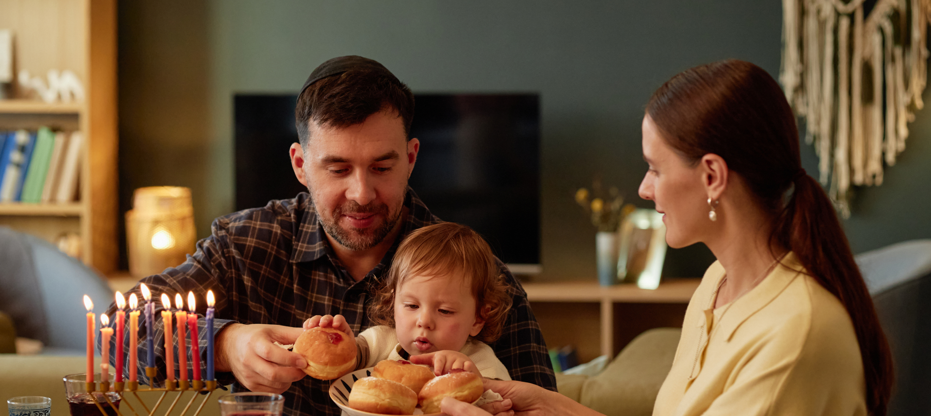 A family of three sits at a table, sharing bread rolls. A menorah with lit candles is on the table, suggesting a Hanukkah celebration. The parents smile and help their young child. The background shows home decor and bookshelves.