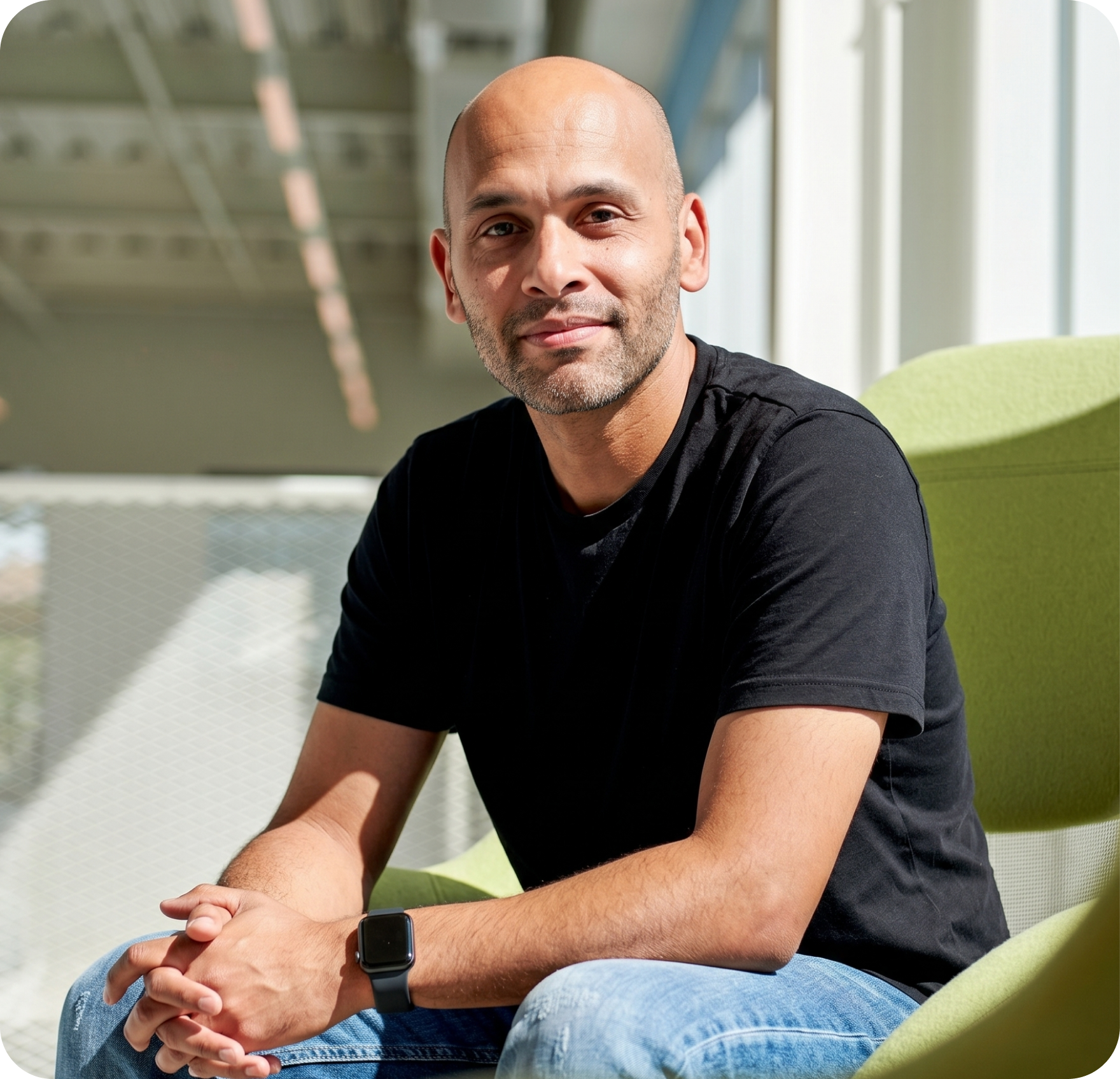 A man with a shaved head wearing a black T-shirt, blue jeans, and a smartwatch sits on a green chair, smiling slightly, in a sunlit modern indoor space.