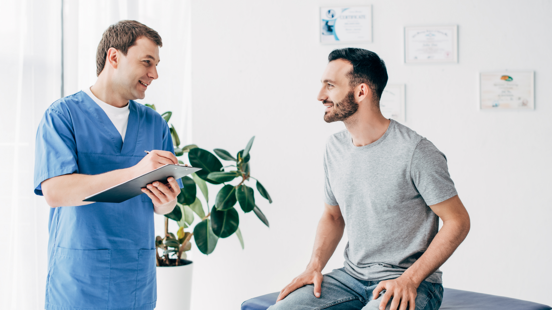 A doctor in blue scrubs smiles and talks with a seated male patient in a grey T-shirt during a consultation in a bright medical surgery with certificates on the wall and a potted plant nearby.