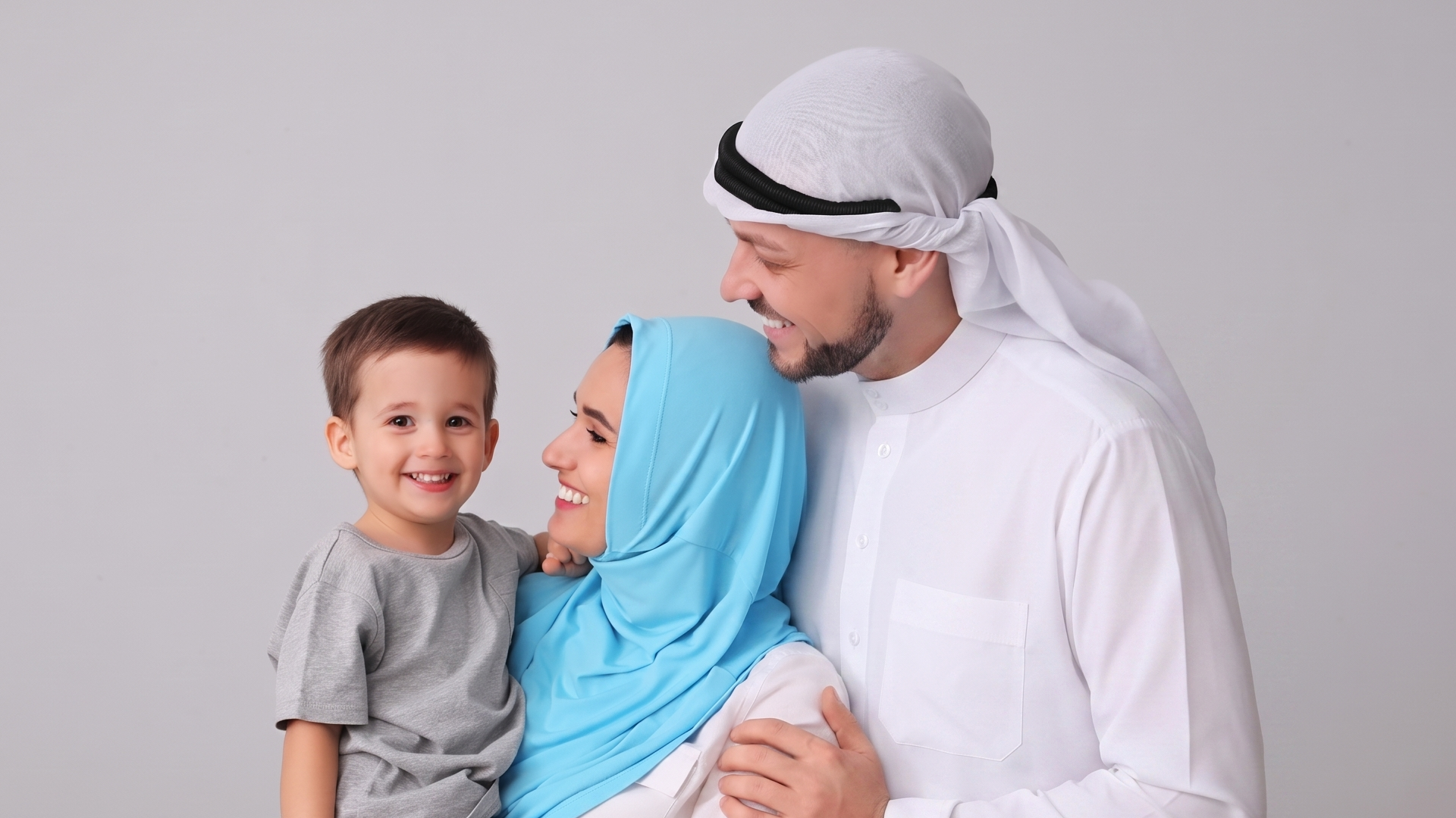 A smiling man in a white kandura and headscarf stands next to a woman in a blue hijab who is holding a young boy. All three are smiling and looking happy against a plain light background.